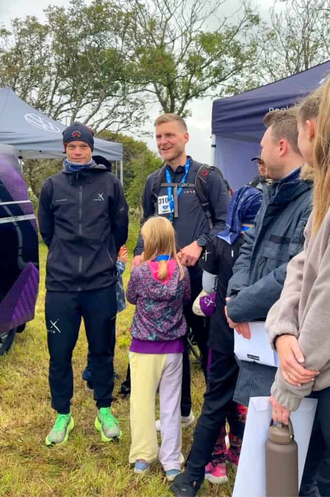 Group of people, including children and adults, gathered outdoors at Markus-løpet 2026 event, engaging in conversation and activities under tents.